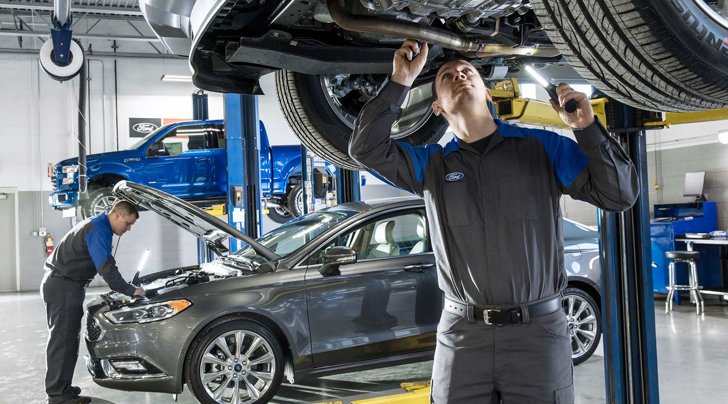 Two Ford technicians work on cars in a busy service bay: one checking under a raised car, another on a sedan's engine.