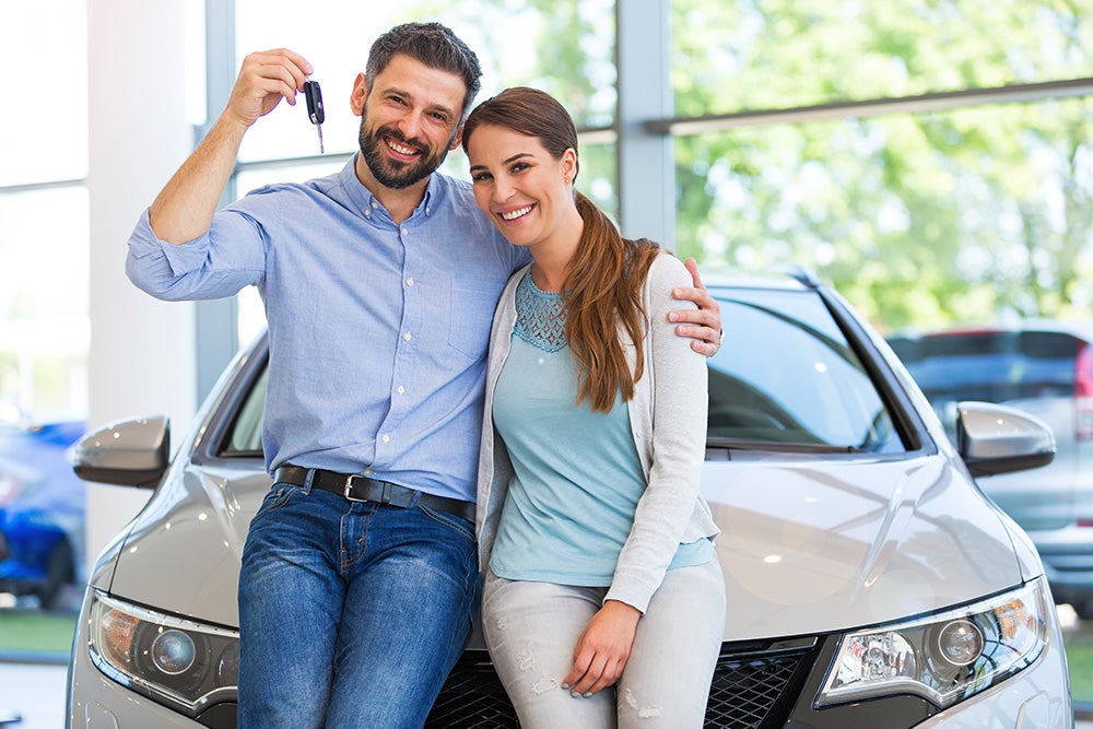 Smiling couple, man holding car keys, leaning on a new silver car in a dealership.